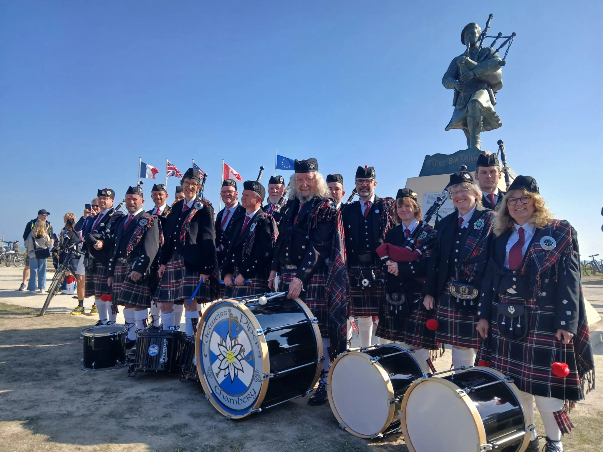 Pipe band posant sous une statue de sonneur de cornemuse, avec des tambours au premier plan.